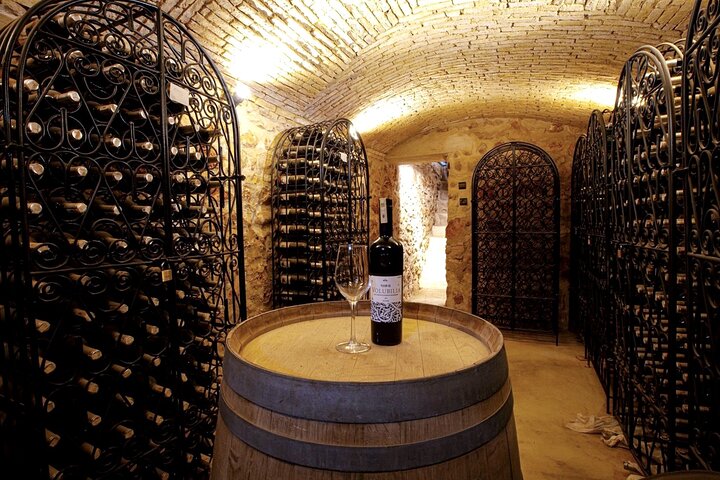 A bottle and a glass on a barrel in a vaulted cellar lined with wine racks