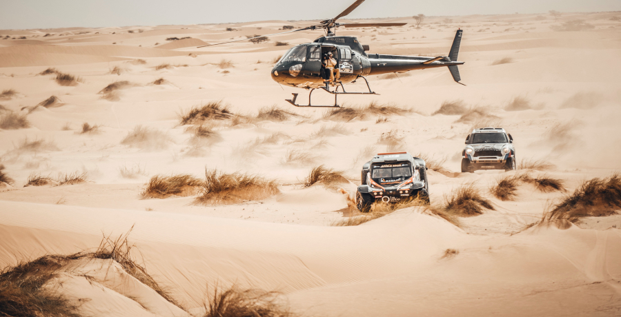 A helicopter flying low over the dunes beside two desert vehicles