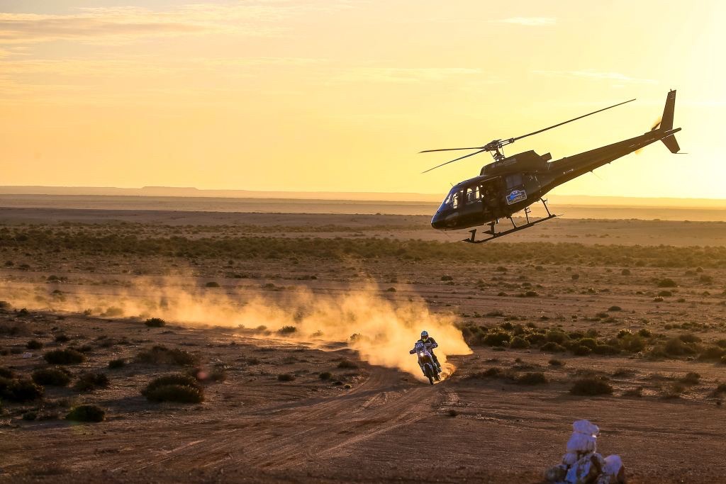 A helicopter above the desert floor at sunset, a trail of dust below